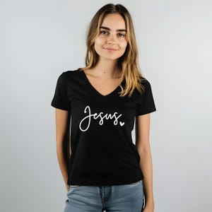 May include: Black V-neck t-shirt with the word "Jesus" in white script. A small white heart is next to the word. The shirt is made of a soft material.