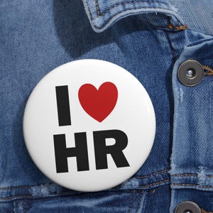 May include: A white button with the text "I ❤️ HR" in black letters and a red heart symbol. The button is pinned to a blue denim jacket.