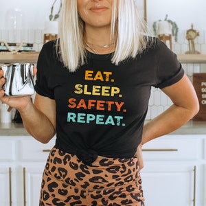 May include: A woman wearing a black t-shirt with the text "EAT. SLEEP. SAFETY. REPEAT." in rainbow colors. She is holding a silver coffee mug and wearing leopard print leggings.