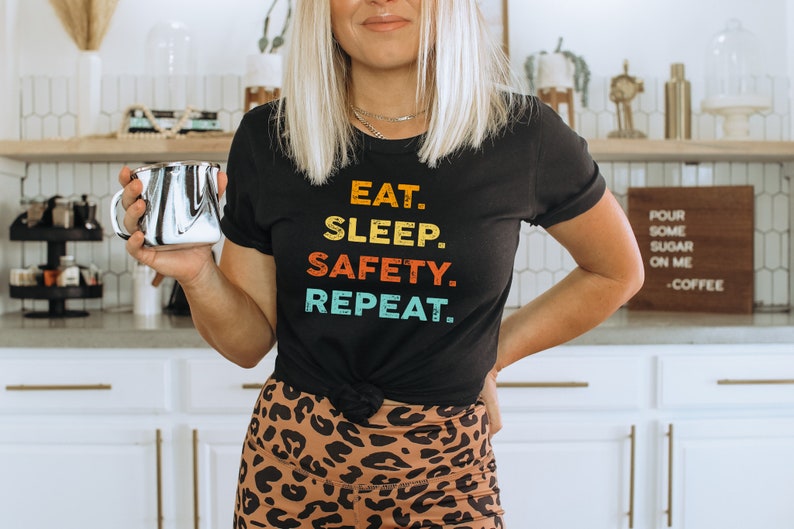 May include: A woman wearing a black t-shirt with the text "EAT. SLEEP. SAFETY. REPEAT." in rainbow colors. She is holding a silver coffee mug and wearing leopard print leggings.