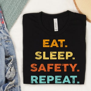 May include: Black t-shirt with a vintage-style graphic that reads "Eat. Sleep. Safety. Repeat."