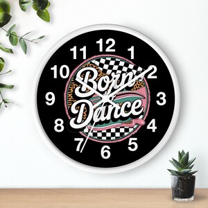 May include: A round wall clock with a white frame and a black face. The clock features the words "Born to Dance" in a retro font, with a pink and leopard print design. The clock hands are silver.