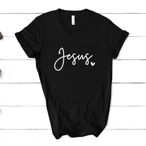 May include: Black V-neck t-shirt with the word "Jesus" in white script, accompanied by a small heart. The shirt is displayed on a white wooden surface.