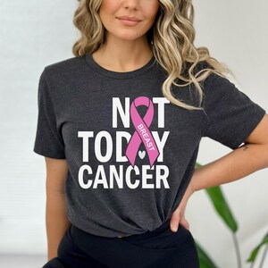 May include: A dark grey t-shirt with the text "NOT TODAY CANCER" in white letters. A pink ribbon with the word "BREAST" is centered on the shirt.