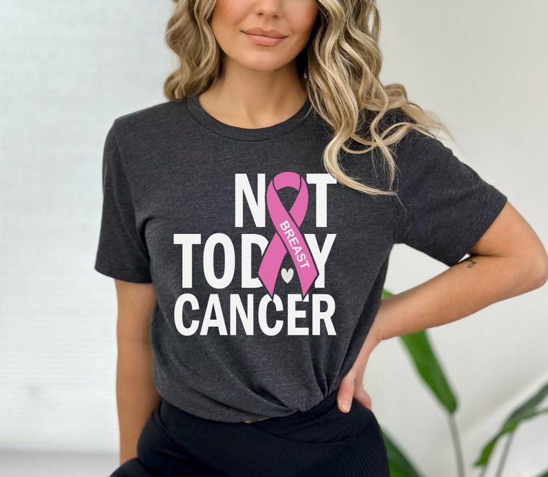 May include: A dark grey t-shirt with the text "NOT TODAY CANCER" in white letters. A pink ribbon with the word "BREAST" is centered on the shirt.