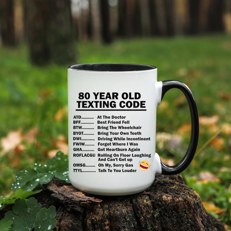May include: White coffee mug with a black handle. The mug has a black rim and the text "80 YEAR OLD TEXTING CODE" in black. Below the text is a list of abbreviations and their meanings, such as "ATD - At The Doctor", "BFF - Best Friend Fell", and "TYL - Talk To You Louder".