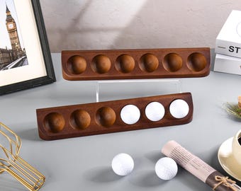 May include: Two wooden golf ball holders with six and five indentations for golf balls. The holders are made of dark brown wood and are stacked on top of each other. There are four white golf balls on the table.