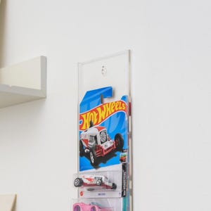 May include: Clear acrylic display case holding several Hot Wheels toy cars. The top car is in a blue and white package with the Hot Wheels logo. Below are two more cars in their original packaging, one white and red, and the other pink.