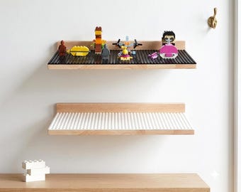 May include: Two light-colored wooden wall shelves with raised back edges. The top shelf features a black building block baseplate surface with various colorful plastic toy figures. The bottom shelf has a white building block baseplate surface.