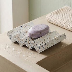 May include: A white soap dish with a multi-colored speckled terrazzo pattern featuring an oval-shaped light purple bar of soap resting in the center. The dish has a distinct zig-zag or accordion-style profile with multiple ridges for drainage.