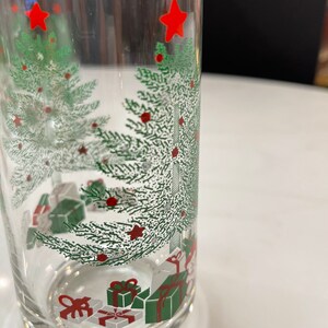 May include: A clear glass tumbler with a Christmas tree design. The tree is green with red ornaments and a red star on top. There are also presents under the tree, wrapped in white and green paper with red bows.