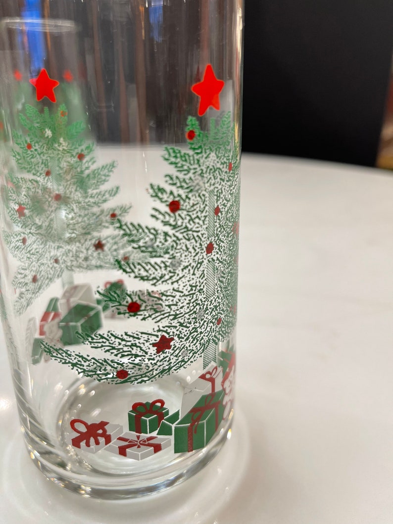May include: A clear glass tumbler with a Christmas tree design. The tree is green with red ornaments and a red star on top. There are also presents under the tree, wrapped in white and green paper with red bows.