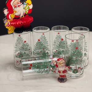 May include: Set of six clear glass tumblers with a Christmas tree design. The glasses feature a green Christmas tree with red stars and presents. A small Santa figurine is in the foreground.