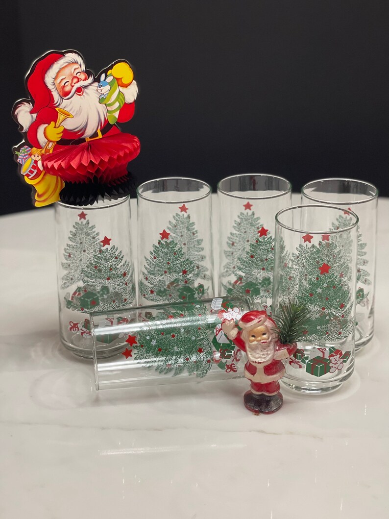 May include: Set of six clear glass tumblers with a Christmas tree design. The glasses feature a green Christmas tree with red stars and presents. A small Santa figurine is in the foreground.