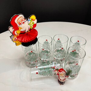 May include: Six clear glass tumblers with a Christmas tree design and red stars. A red and white Santa Claus paper fan decoration is on top of one of the glasses. A small Santa Claus figurine is on the table.