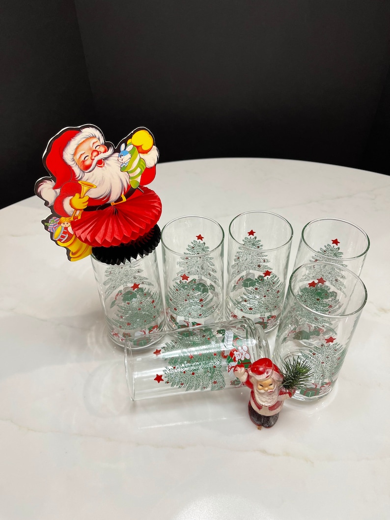 May include: Six clear glass tumblers with a Christmas tree design and red stars. A red and white Santa Claus paper fan decoration is on top of one of the glasses. A small Santa Claus figurine is on the table.