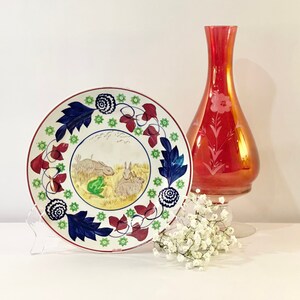 May include: A white ceramic plate with a blue, green, and red floral design and a scene of two rabbits in a field. The plate is on a white stand. A red glass vase with a floral design is in the background.