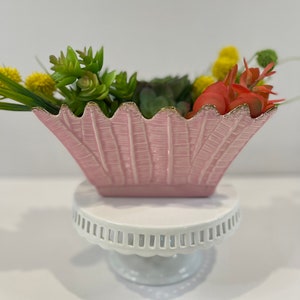 May include: A pink ceramic planter with a scalloped rim and gold trim. The planter is filled with green succulents and red and yellow flowers. The planter is sitting on a white cake stand.