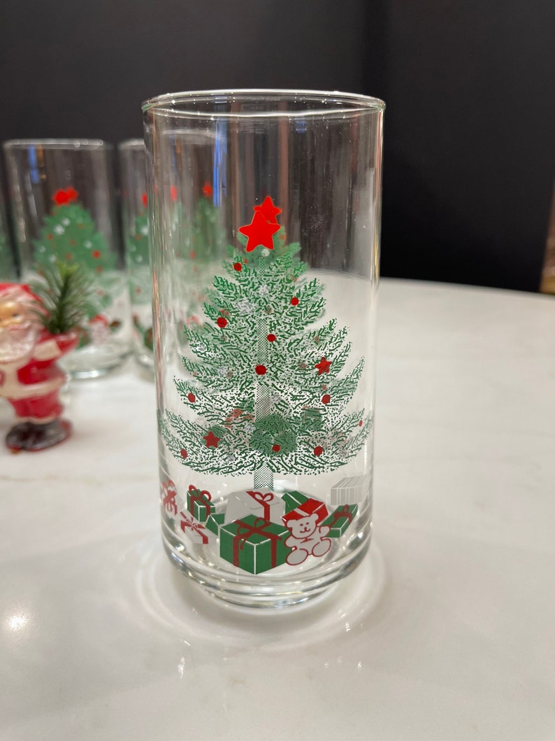 May include: Set of six clear glass tumblers with a Christmas tree design. The tree is green with red ornaments and a red star on top. There are presents and a teddy bear at the base of the tree.