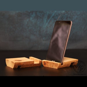Può includere: Un supporto per telefono in legno con uno smartphone grigio scuro appoggiato sul supporto. Il supporto è in legno naturale con una finitura rustica. Il telefono è uno smartphone moderno con uno schermo nero e una cornice argentata. Il supporto è un design semplice con un'unica fessura per il telefono. Il supporto è un ottimo modo per mantenere il telefono in posizione verticale e facilmente accessibile.
