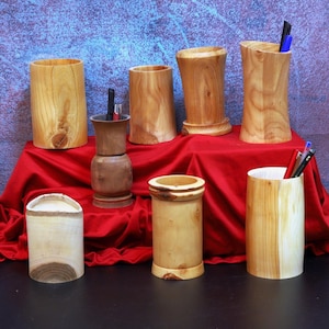 May include: A selection of handcrafted wooden desk tidies and pen holders. The items vary in shape and size, displaying natural wood grain patterns. Some holders contain pens and pencils, and are displayed on a red cloth.