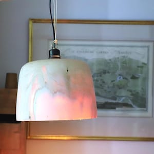 Könnte beinhalten: Eine Holz-Pendelleuchte mit einem glockenförmigen Schirm. Der Schirm hat ein natürliches Holzmaserungsmuster mit rosa und weißen Farbtönen. Das Licht hängt an einem schwarzen Kabel und einer Metallhalterung.