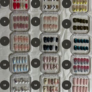 Op de afbeelding: Vijftien kleine transparante doosjes, elk met een set van tien kunstnagels met verschillende ontwerpen. De ontwerpen omvatten koe-print, glitter, ombre en abstracte patronen.