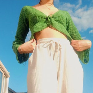 Op de afbeelding: Een groen gehaakt crop top met een strik aan de voorkant en lange mouwen. Het topje wordt gedragen over een witte, wijde broek met een trekkoord.