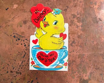 Op de afbeelding: Een vintage Valentijnskaart met een geel kuiken in een blauwe theekop. Het kuiken houdt een rood hart vast en de kaart zegt "Hi Chucky! You're my Cup O' Tea".