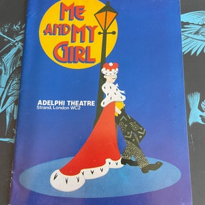 May include: A vintage playbill for the musical "Me and My Girl" at the Adelphi Theatre in London. The playbill features a cartoon illustration of a couple in period dress, with a yellow street lamp and a blue background.