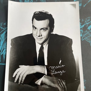 Pode incluir: Fotografia em preto e branco de Mario Lanza, um famoso tenor, vestindo um terno e gravata. Ele está sorrindo e olhando para a câmera. A foto está assinada "Mario Lanza".