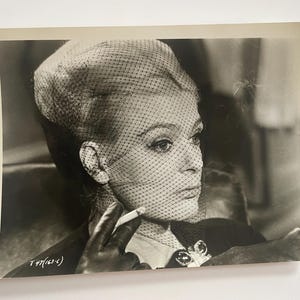 Pode incluir: Uma fotografia a preto e branco de uma mulher usando um véu e um vestido escuro. Ela está segurando um cigarro na mão direita e olhando para a direita. A foto foi tirada de um ângulo baixo.