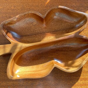 May include: A wooden serving dish shaped like a leaf with two separate compartments. The dish is made of light brown wood and has a smooth, polished finish.