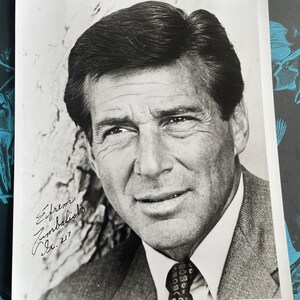 May include: Black and white photo of a man wearing a suit and tie. He is smiling and looking at the camera. The photo is signed "Efrem Zimbalist Jr. 2-17".