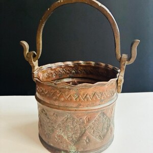 May include: A cylindrical copper bucket featuring a scalloped rim, a swinging metal handle, and embossed diamond patterns containing floral motifs. The exterior surface displays a weathered patina with visible green oxidation and metallic texture.