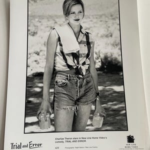 May include: A monochrome promotional image for the film "Trial and Error" starring Charlize Theron. She is dressed in a tied plaid shirt and denim shorts, holding two jars. A white towel is draped over her shoulder.