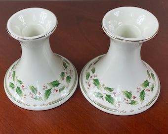 May include: Two white ceramic candle holders with a green and red holly design. The holders have a silver rim around the top.