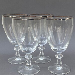 May include: Set of four clear glass wine glasses with a silver rim. The glasses have a classic design with a tall, slender bowl and a short stem.