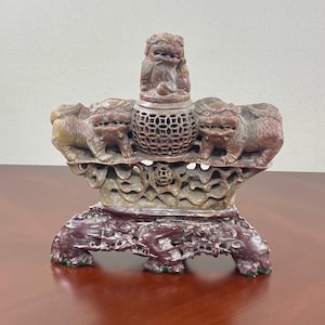 May include: A carved stone incense burner with three lion figures and a decorative base. The burner is made of red and brown stone and has a intricate design.