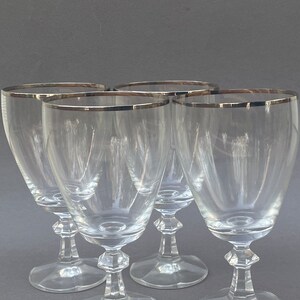 May include: Set of four clear glass wine glasses with a silver rim. The glasses have a classic design with a tall, slender bowl and a short stem.