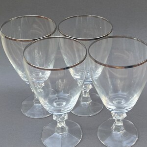 May include: Set of four clear glass wine glasses with a silver rim. The glasses have a tall, slender shape with a round bowl and a stem.