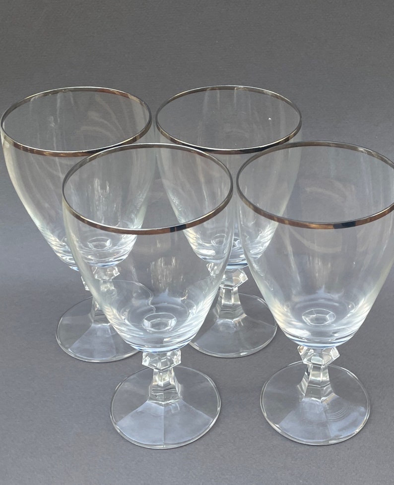 May include: Set of four clear glass wine glasses with a silver rim. The glasses have a tall, slender shape with a round bowl and a stem.