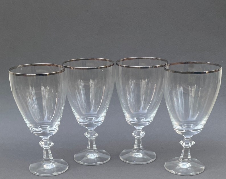 May include: Four clear glass wine glasses with silver rims. The glasses have a traditional shape with a wide bowl and a long stem.
