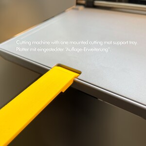 May include: A close-up of a gray cutting machine with a yellow extension tray. The text "Cutting machine with one mounted cutting mat support tray. Plotter mit eingesteckter "Auflage-Erweiterung"." is visible on the machine.