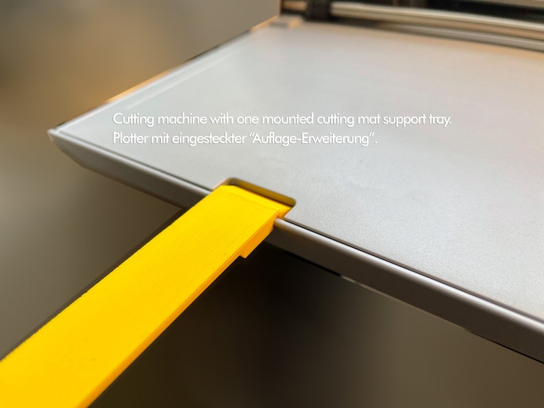 May include: A close-up of a gray cutting machine with a yellow extension tray. The text "Cutting machine with one mounted cutting mat support tray. Plotter mit eingesteckter "Auflage-Erweiterung"." is visible on the machine.