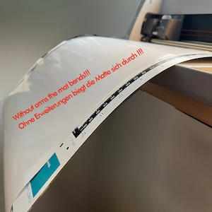 May include: A white cutting mat is being fed into a cutting machine. The mat is bending and the text "Without arms the mat bends!!! Ohne Erweiterungen biegt die Matte sich durch!!!" is visible on the mat.