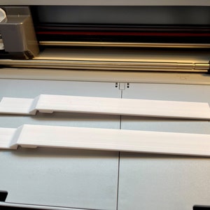 May include: Two white plastic strips, possibly for a craft project, are lying on a gray surface inside a cutting machine.