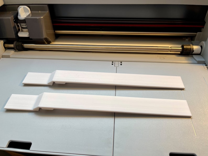 May include: Two white plastic strips, possibly for a craft project, are lying on a gray surface inside a cutting machine.