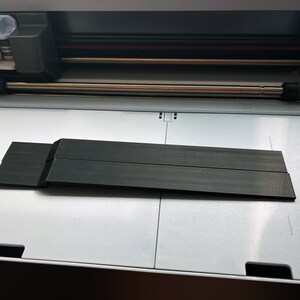 May include: A black plastic cutting mat for a craft cutting machine. The mat is placed on the cutting bed of the machine.