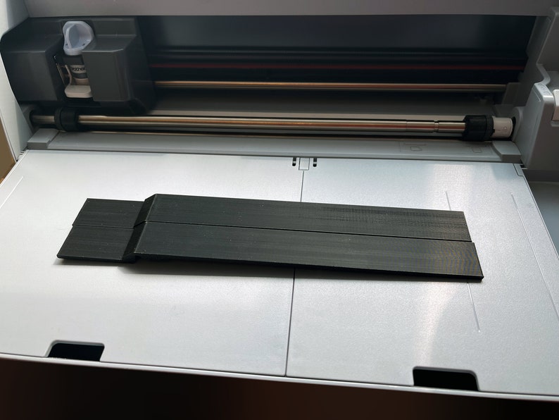 May include: A black plastic cutting mat for a craft cutting machine. The mat is placed on the cutting bed of the machine.